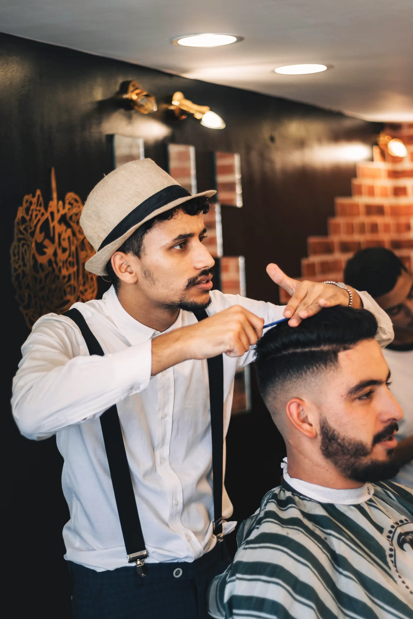 Barbero con sombrero y tirantes corta el cabello a un cliente frente a un muro de ladrillo