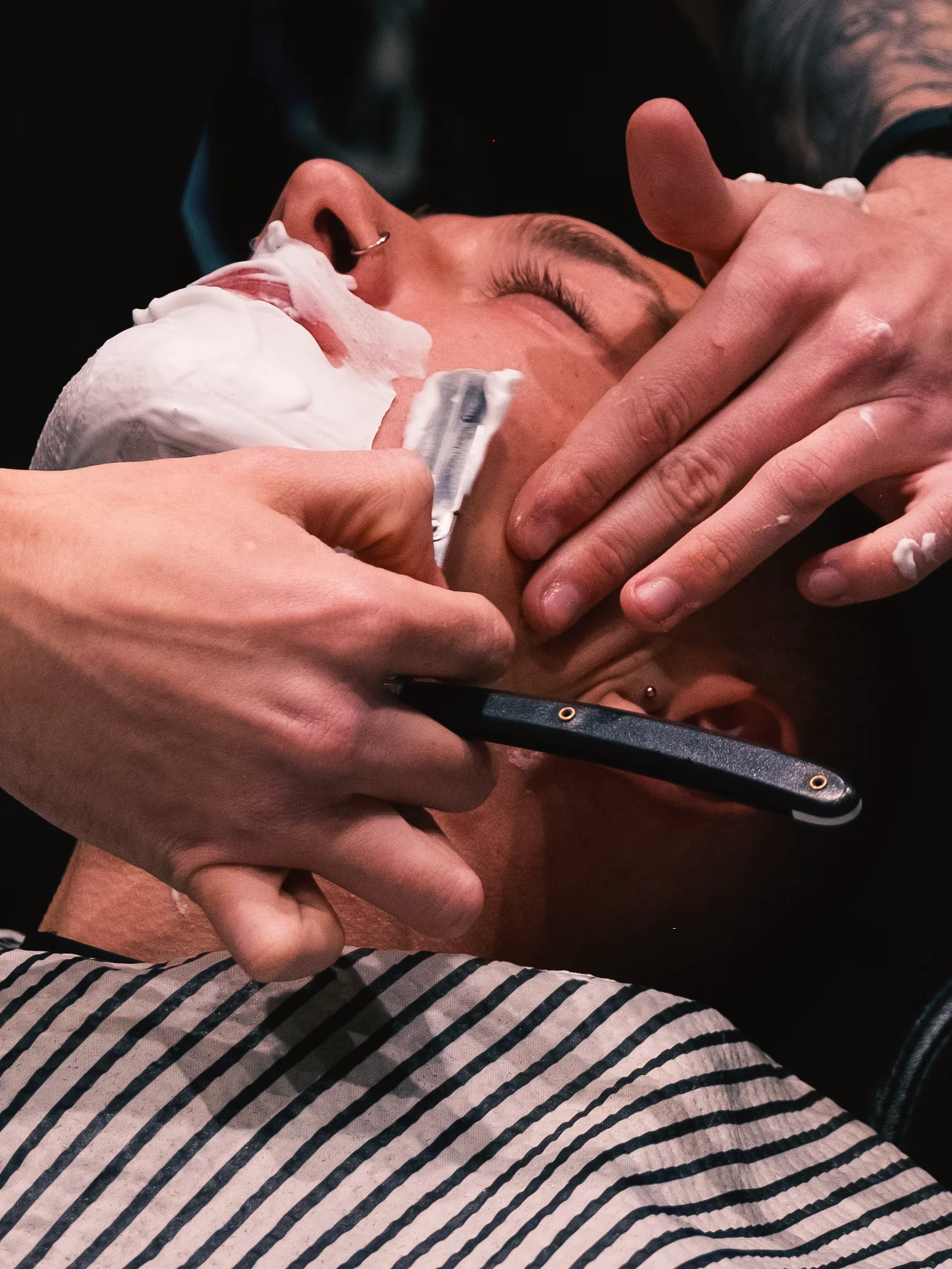 Barbero con brazos tatuados afeita con navaja a un cliente con espuma en la cara